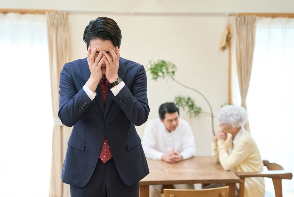 A man in a suit covering his face with his hands while his parents sit at a table behind him