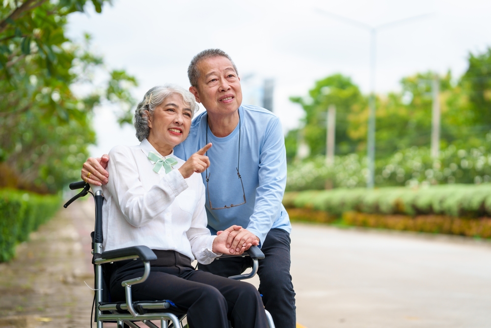 An older couple outdoors, the husband pointing ahead while his wife holds his hand from her wheelchair