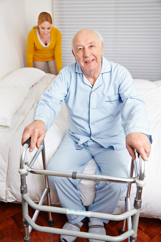 An older man in pajamas standing up from a bed with a walker, a nurse behind him