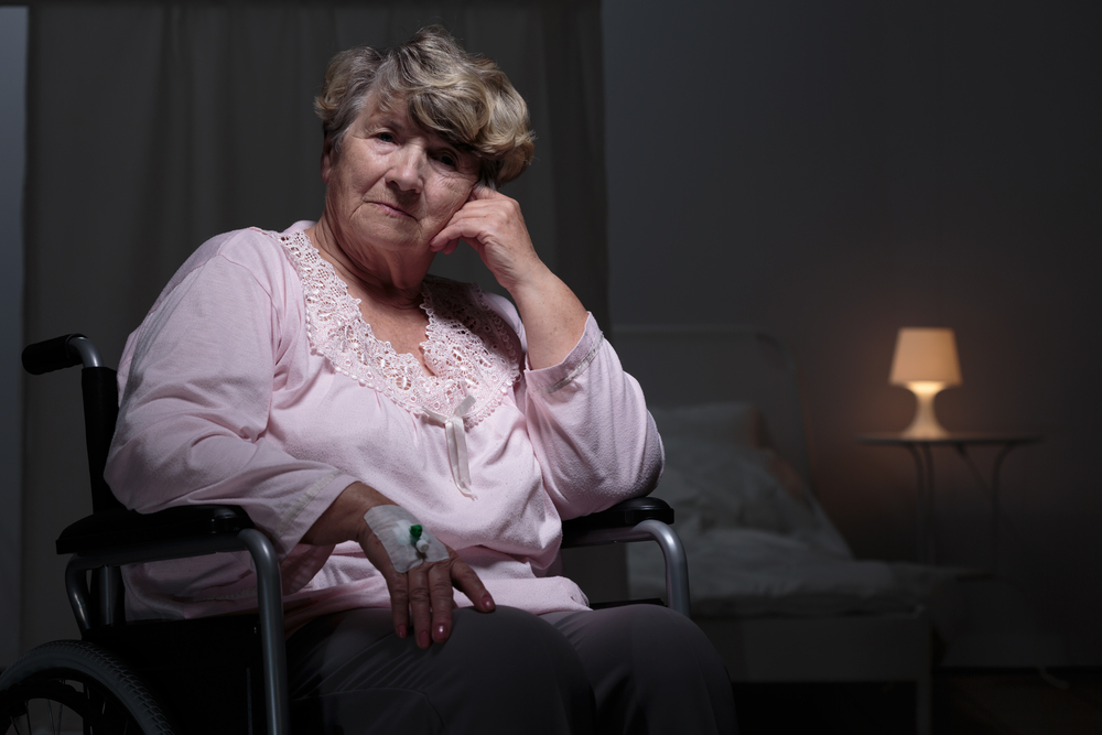An older woman sitting in a wheelchair in a dim bedroom, an IV line in her arm