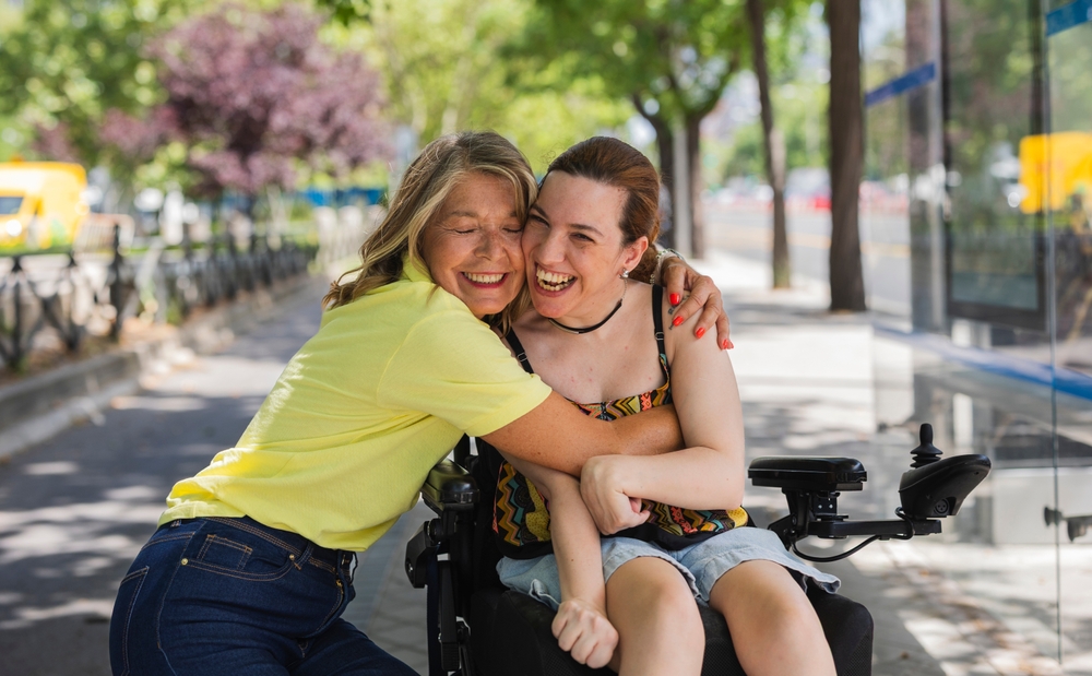 A mother hugging her laughing adult daughter, who uses a power wheelchair, on a city sidewalk