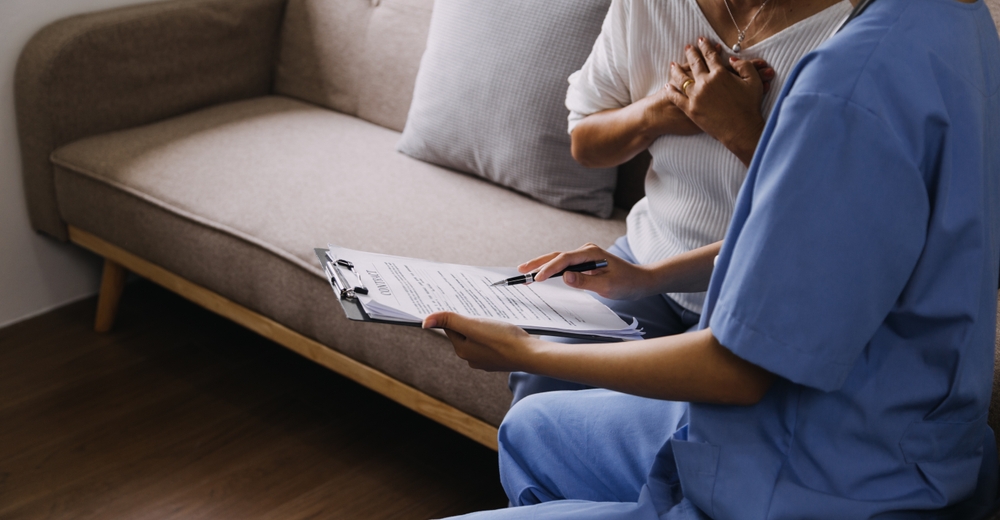 A nurse filling in a form on a clipboard while sitting with a patient at home