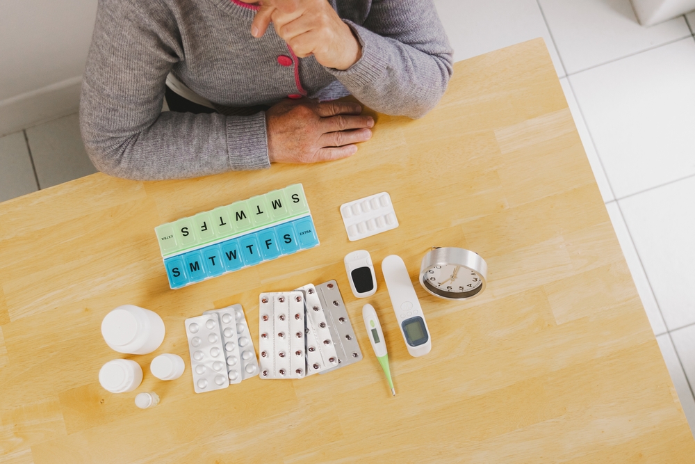 An older person at a table with a weekly pillbox, blister packs, a thermometer and a glucose meter