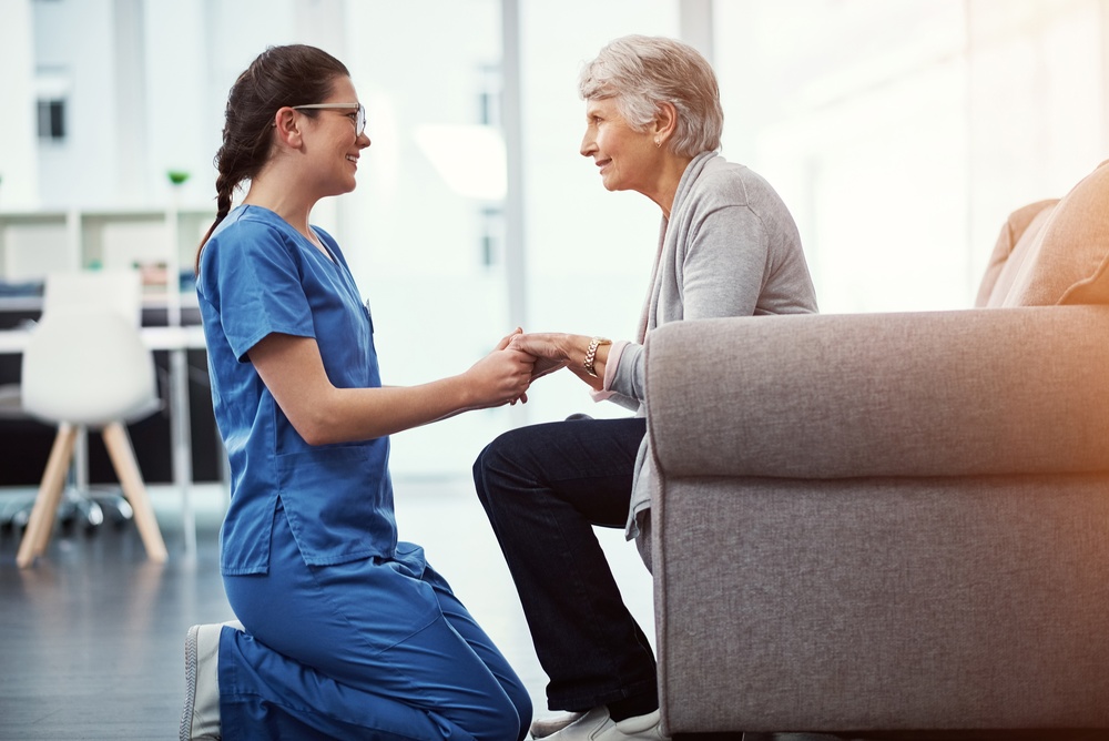 A home health aide in scrubs kneeling to hold an older woman's hands