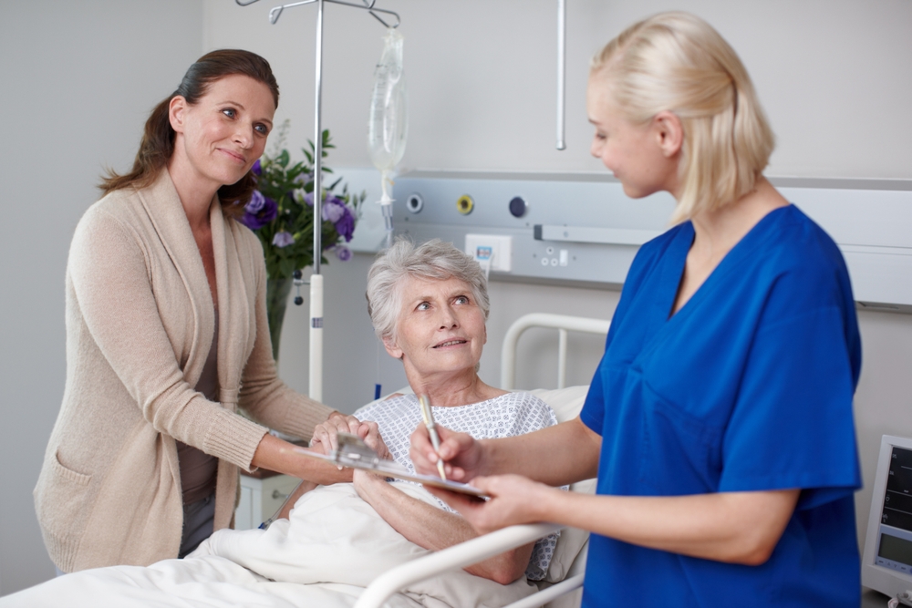A nurse going over discharge papers with an older patient in a hospital bed while her daughter listens