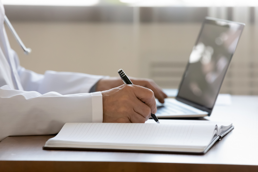 A doctor writing in a notebook beside a laptop