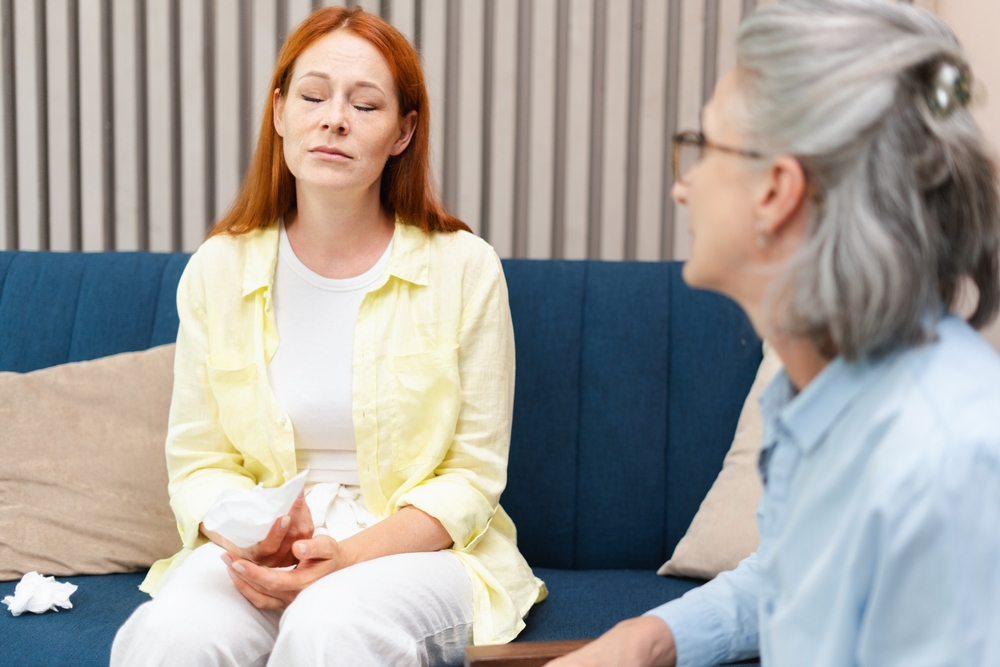 A young woman with her eyes closed on a couch, a therapist listening beside her