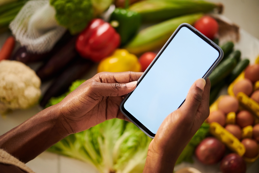 A hand holding a phone over a basket of fresh vegetables