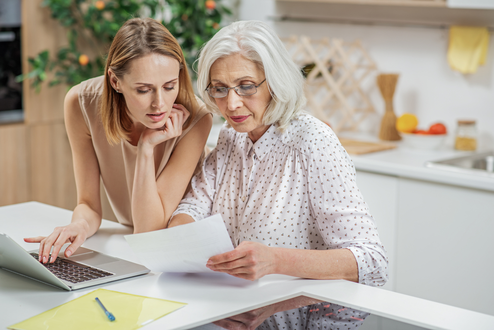 A daughter and her elderly mother reviewing a document at a kitchen table with a laptop