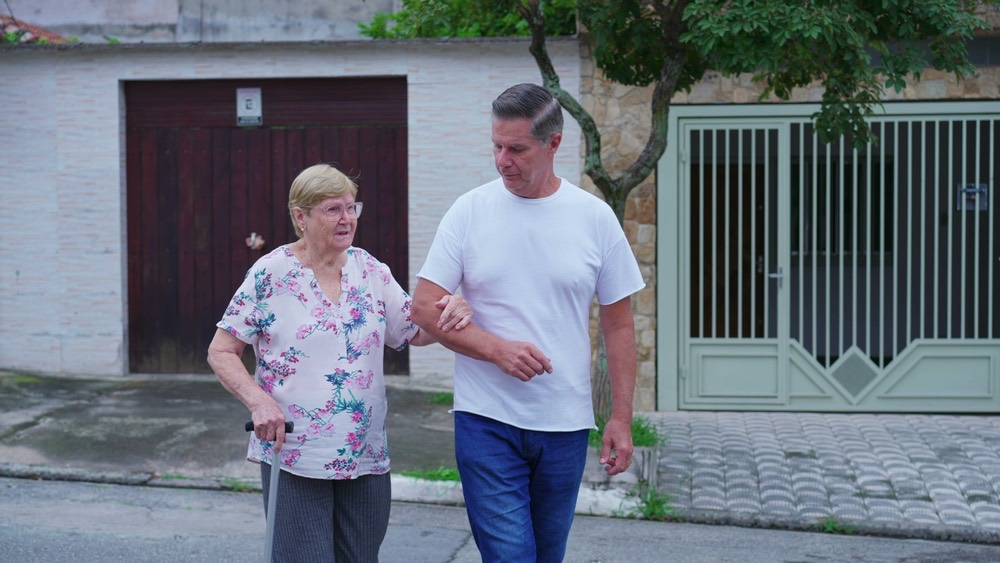 An adult son walking arm in arm with his mother, who uses a cane