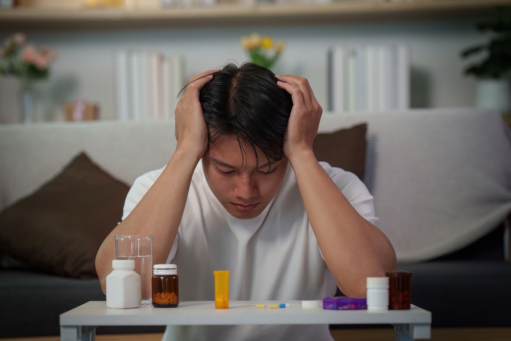 A young man with his head in his hands in front of a table of pill bottles