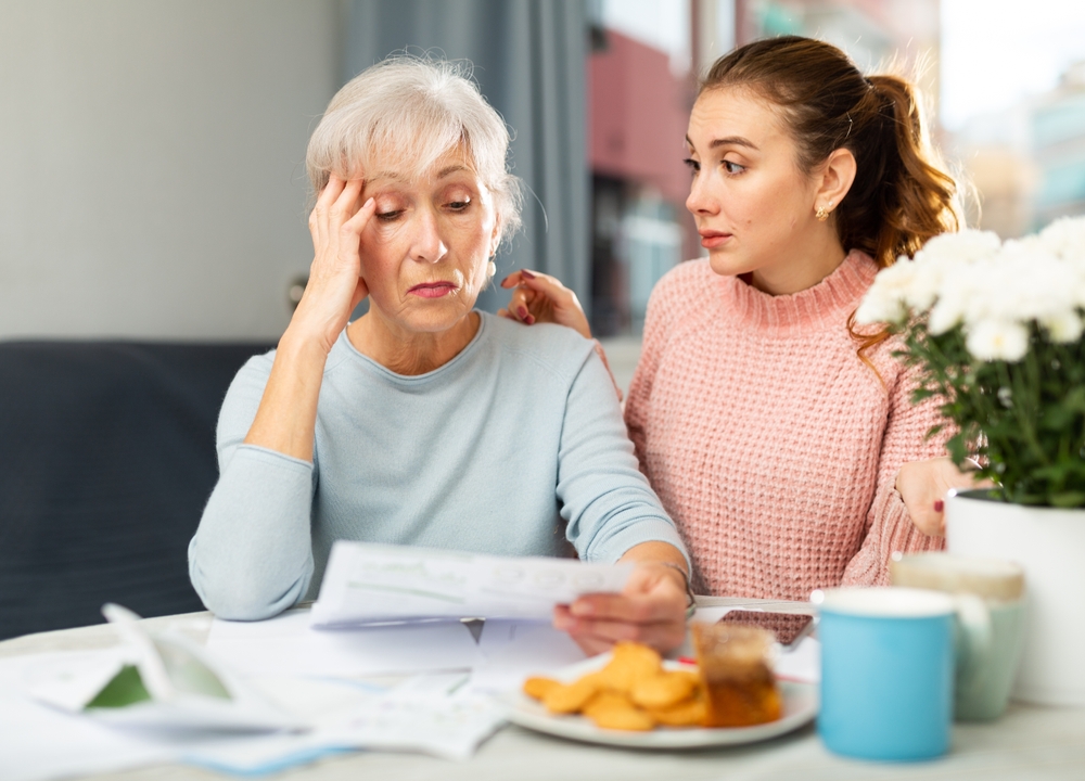An elderly woman holding her head over a pile of bills while her daughter comforts her