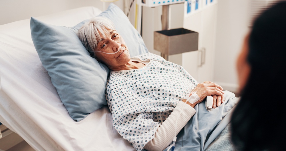 An older woman in a hospital bed with an oxygen line, a visitor at her side