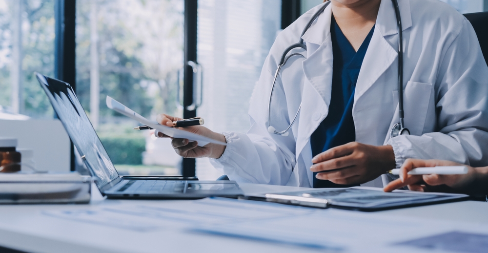 A doctor at a desk handing over a document beside a laptop and a tablet